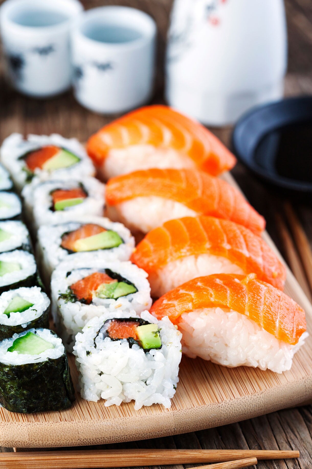 Fresh salmon nigiri sushi and assorted sushi rolls arranged on a bamboo serving board with chopsticks, traditional Japanese sake cups, and soy sauce dish in the background.