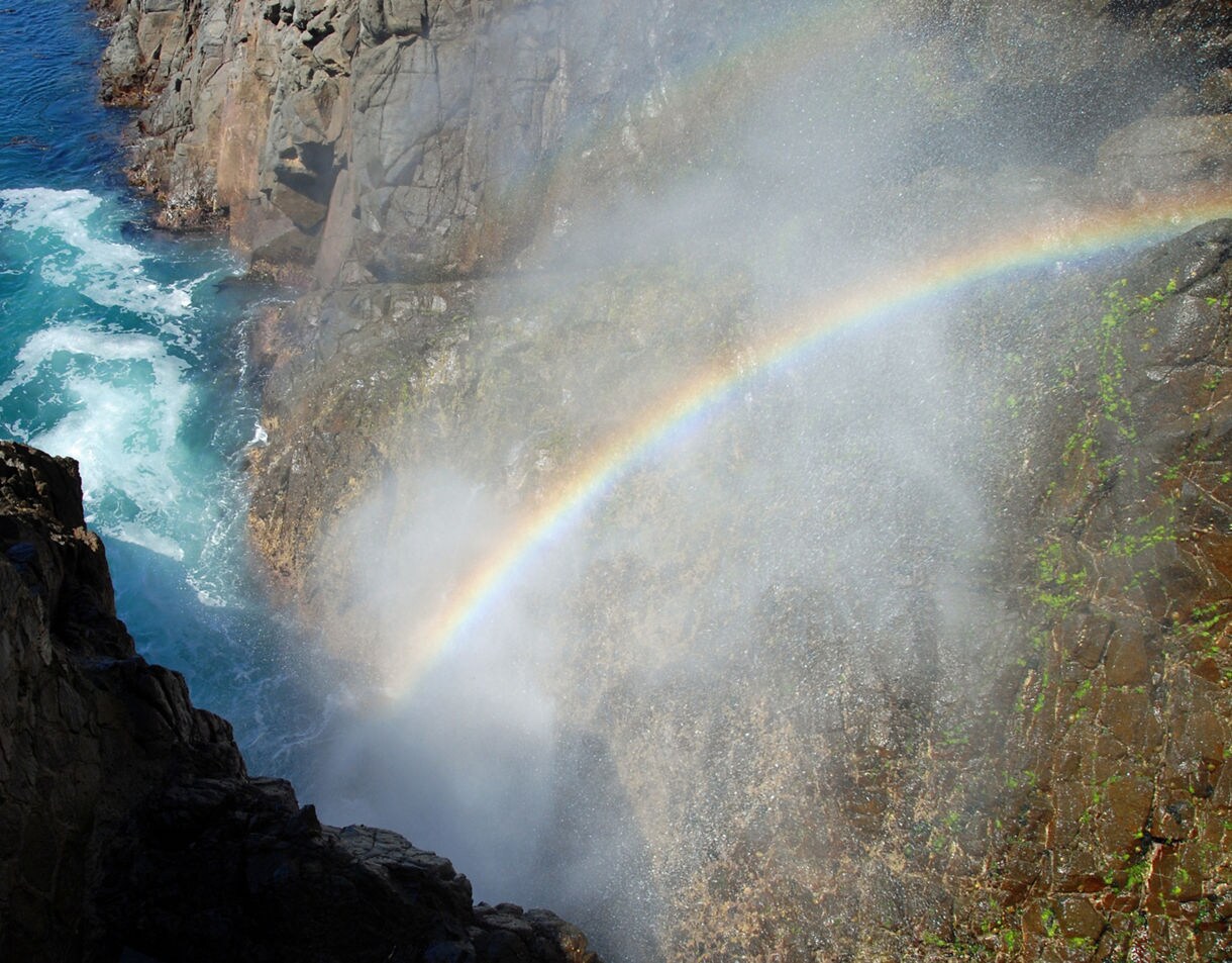 Ocean waves surge into the rocky cliffs at La Bufadora blowhole in Baja California, with mist forming a vivid rainbow over the blue water.