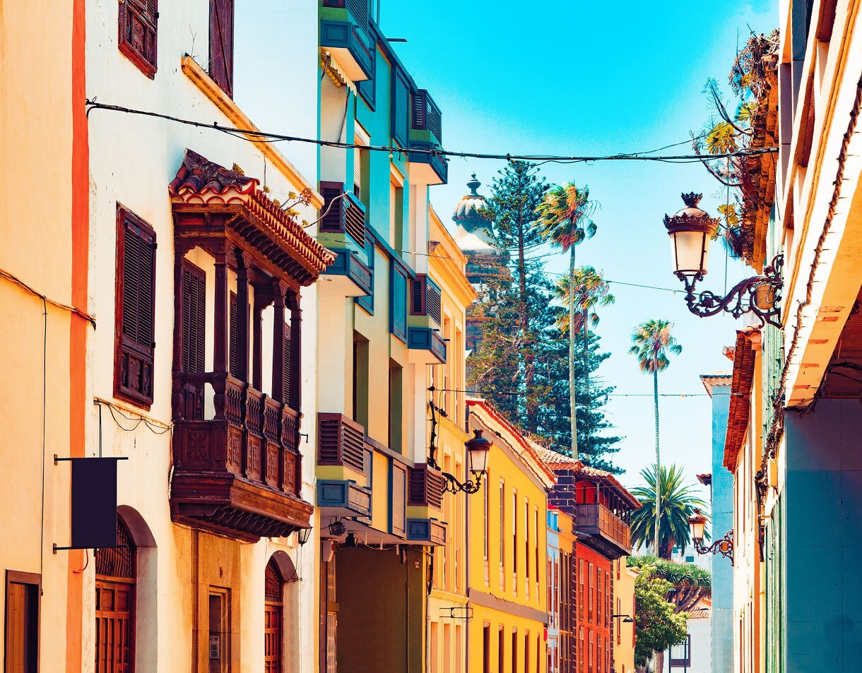 You can walk La Laguna’s colorful streets that are lined with colonial-era homes and churches. Alt: Vibrant historic street in La Laguna lined with colorful houses, wooden balconies and palm trees in the background.