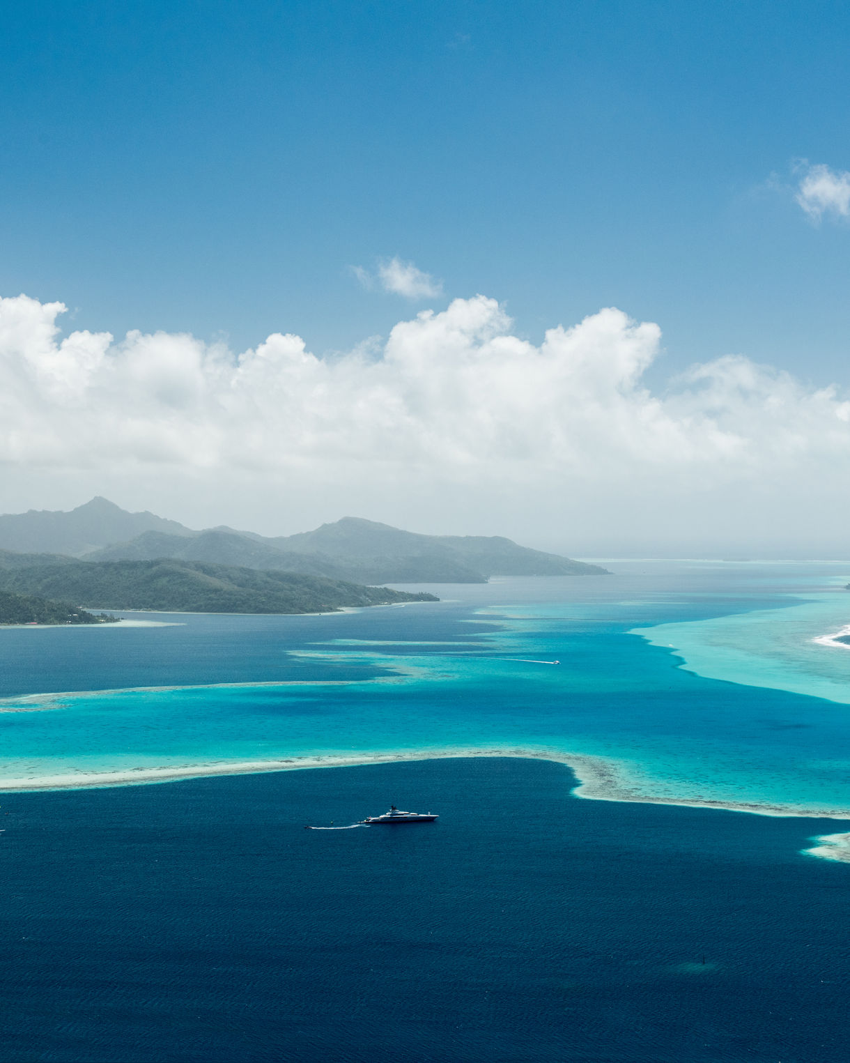 Aerial view of Raiatea’s lagoon showing deep blue ocean, turquoise shallows and green mountains under a bright tropical sky.