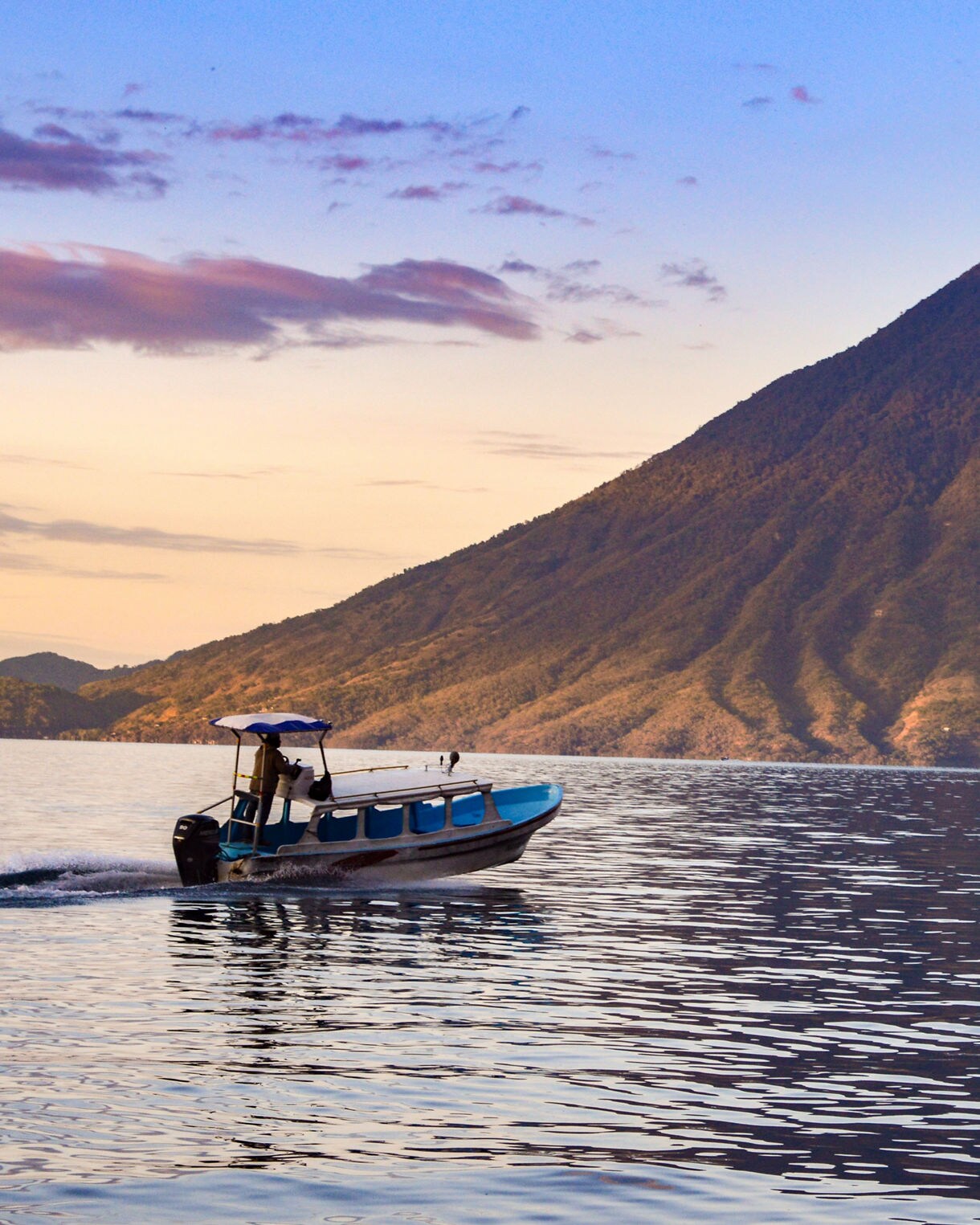 A small boat cruising across Lake Atitlán in Guatemala at sunset, with calm waters reflecting the light and a large volcanic mountain rising in the background.