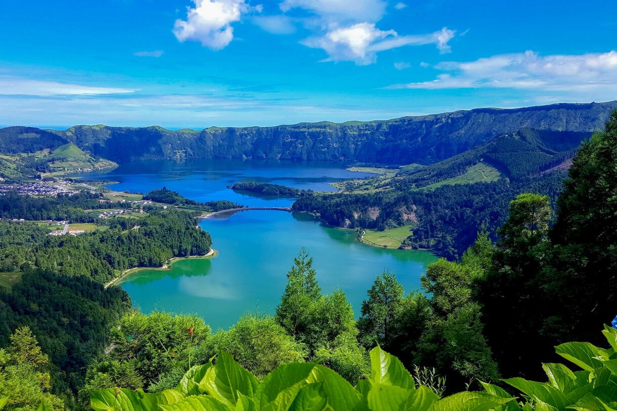 Lake Azul volcanic crater lake on the island of São Miguel, Azores, Portugal, viewed from an elevated vantage point, surrounded by lush green forests and steep caldera walls, with a small coastal town visible in the distance under a bright blue sky with scattered clouds.