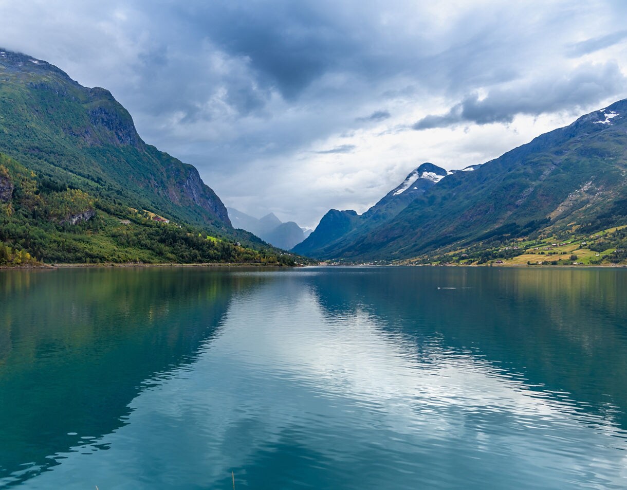 Lake Lovatnet in Norway with still turquoise water reflecting surrounding green mountains and cloudy skies.