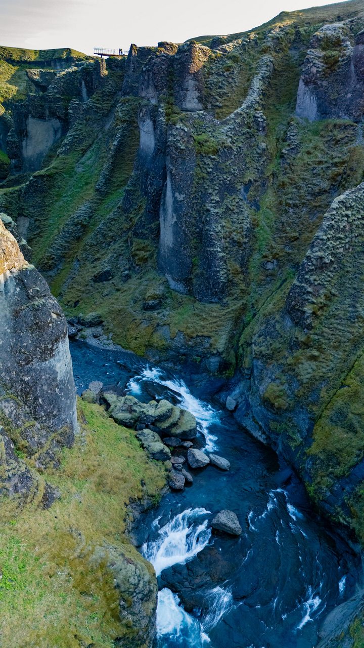 Narrow river with whitewater flowing between tall mossy canyon walls under soft daylight.