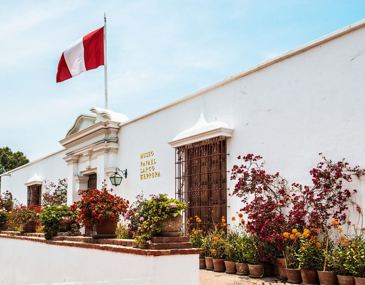 White colonial-style exterior of the Larco Museum in Lima with a Peruvian flag flying above and colorful flowers lining the entrance.