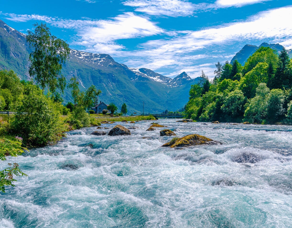 Rapid glacial river at Laukifossen in Norway, with foaming turquoise water surrounded by green trees and snow-capped peaks in the distance.