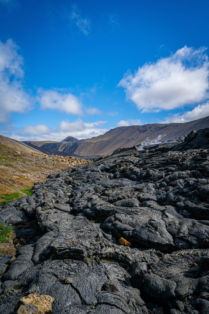 Dark, hardened lava flows stretch across a rocky valley with low mountains and scattered clouds in the background