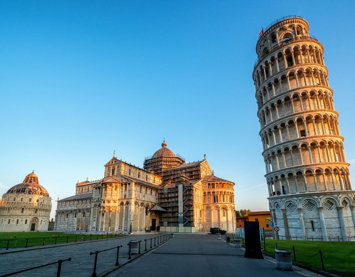 The Leaning Tower of Pisa and the Pisa Cathedral at golden hour, both illuminated by warm sunlight against a clear blue sky on a spacious open plaza.