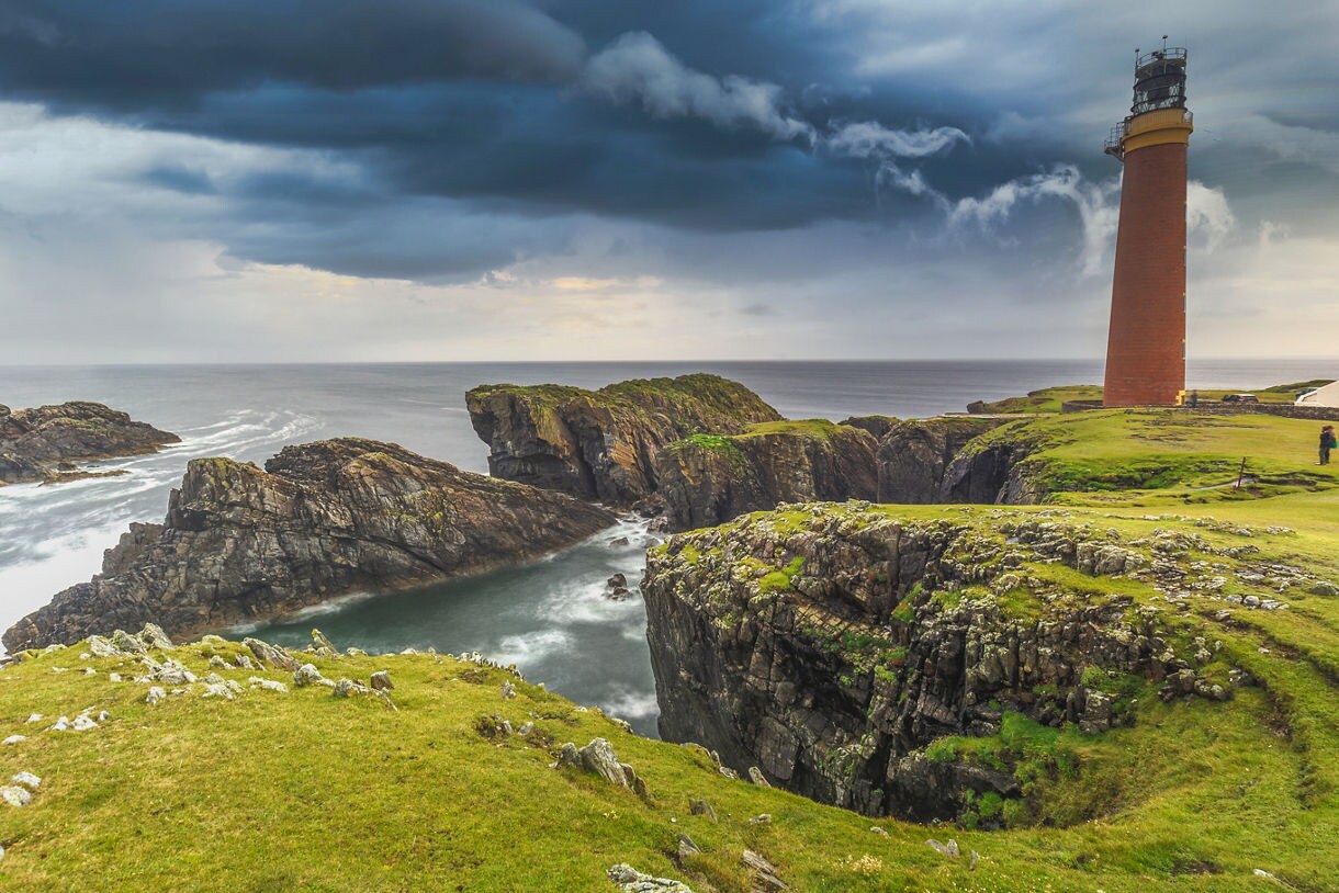 Coastal cliffs on the Isle of Lewis with a tall brick lighthouse overlooking rugged rock formations and the Atlantic Ocean beneath dark, swirling storm clouds.