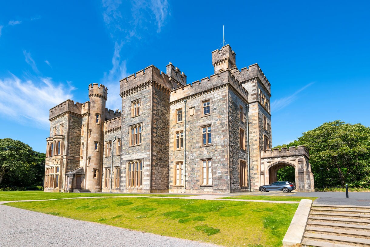Exterior view of Lews Castle on the Isle of Lewis, showcasing its stone towers, arched entryway, bright green lawns and a clear blue sky overhead.