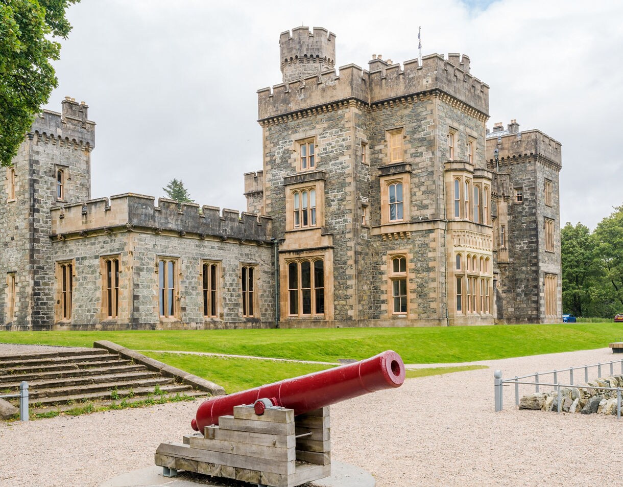 Stone-built Lews Castle in Stornoway with crenellated towers, tall windows and a red cannon displayed on the lawn, surrounded by green trees under an overcast sky.