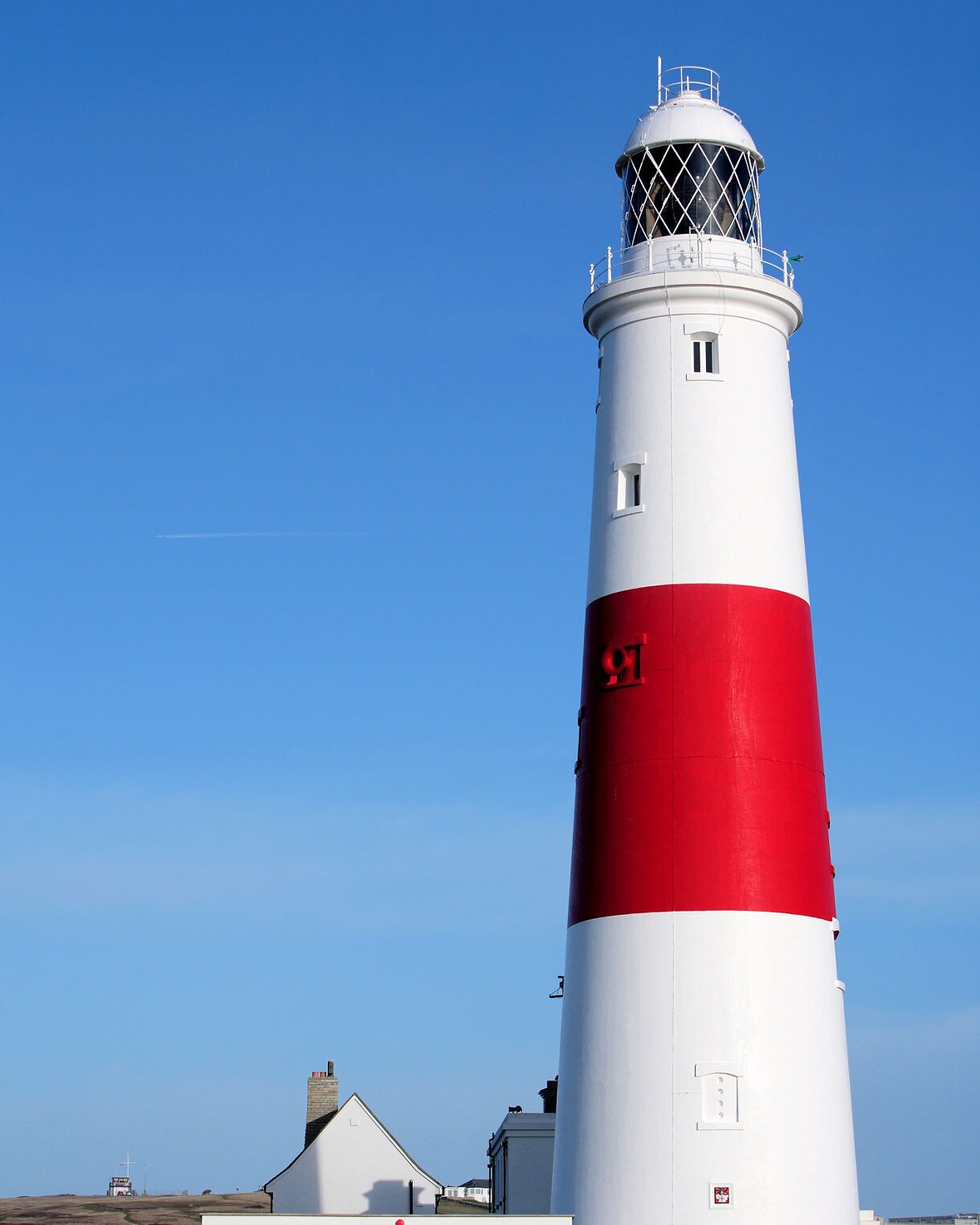 Tall white lighthouse with a wide red band near the middle, set against a clear blue sky with small buildings at its base.