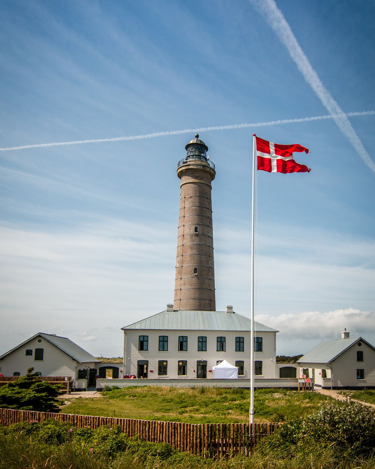 The Grey Lighthouse at Grenen Beach, Denmark, with a Danish flag waving in front under streaked blue skies.