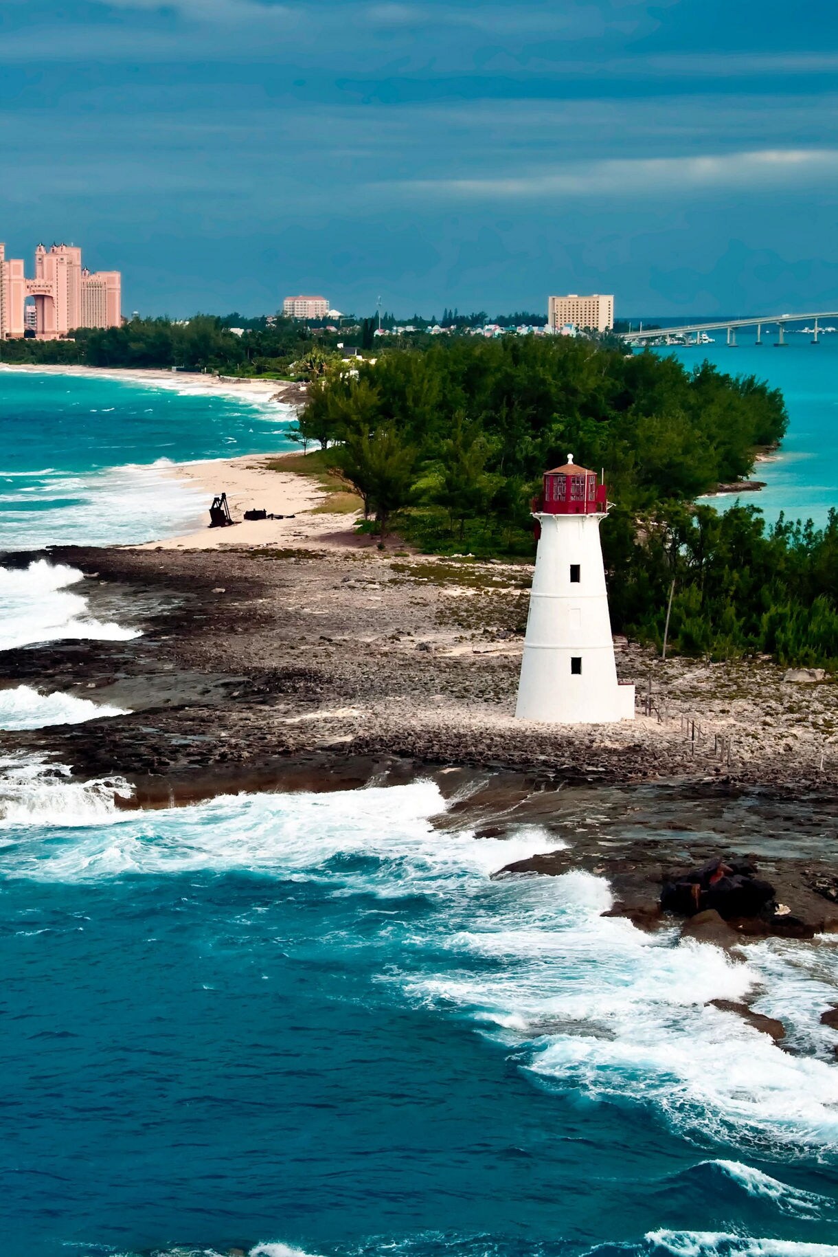 Nassau lighthouse on a rocky coastline with waves crashing, and the Atlantis resort buildings visible in the background across turquoise waters.