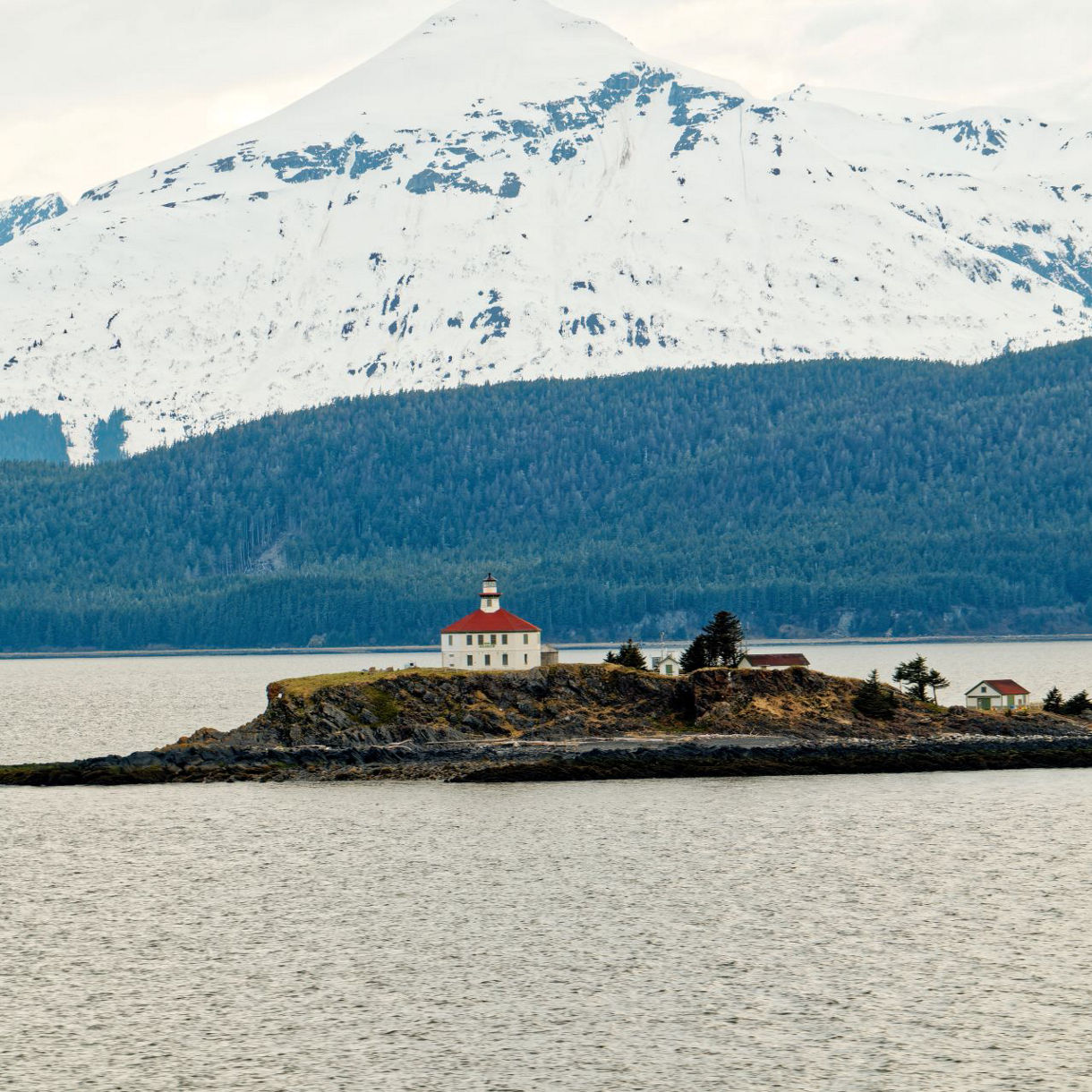 Eldred Rock Lighthouse on a small island near Skagway, Alaska, surrounded by calm waters with towering snow-covered mountains and lush forests in the distance.