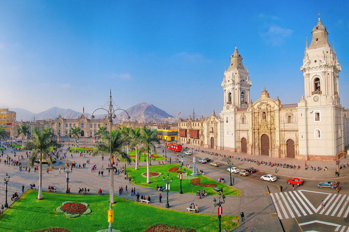 Panoramic view of Lima’s Plaza Mayor with bright yellow colonial buildings, a grand cathedral, green gardens and crowds moving through the historic square under a clear blue sky.