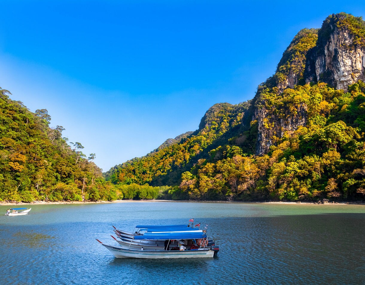 Long-tail boats floating on a calm river bordered by steep limestone hills covered in dense tropical forest under a bright blue sky.