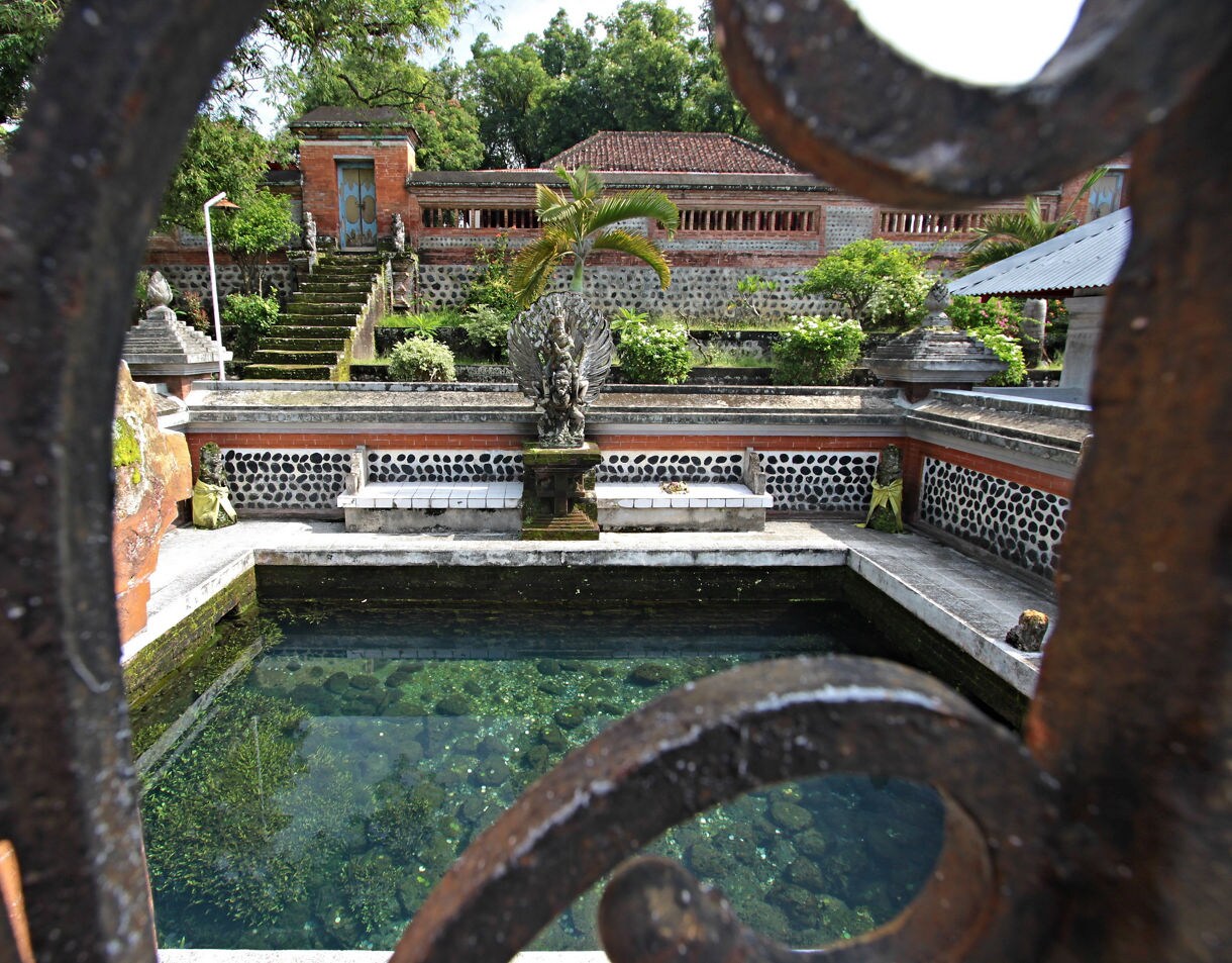 View through an ornate gate of Lingsar Temple’s inner courtyard in Lombok, showing a clear spring pool, stone benches, lush greenery and a central carved statue framed by moss-covered steps and red-brick walls.