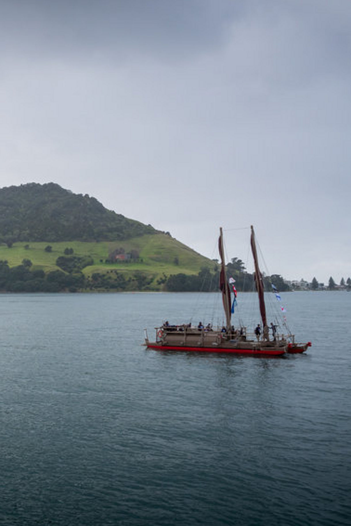 A traditional double-hulled Waka sails across calm waters of Tauranga, with Mt. Maunganui in the background.