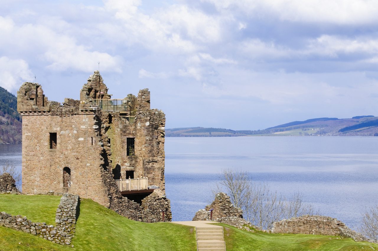 Image of castle ruins with a lake in the background, featuring a stone structure with a wooden balcony, a grassy area, and a stone staircase leading to the water's edge.