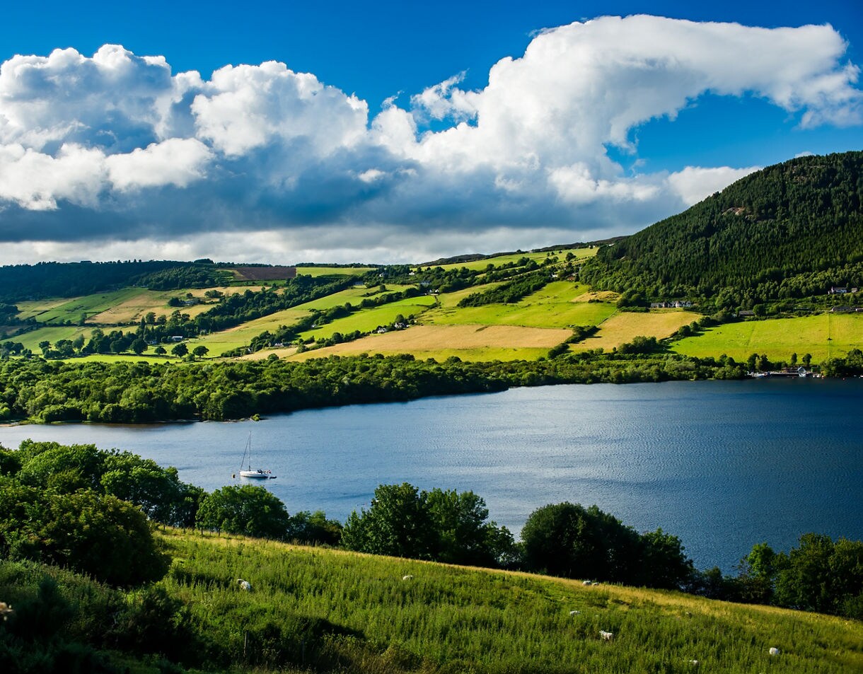 Scenic view of Loch Ness in Scotland with lush green hills, patchwork fields and a sailboat on the calm blue water beneath large clouds.