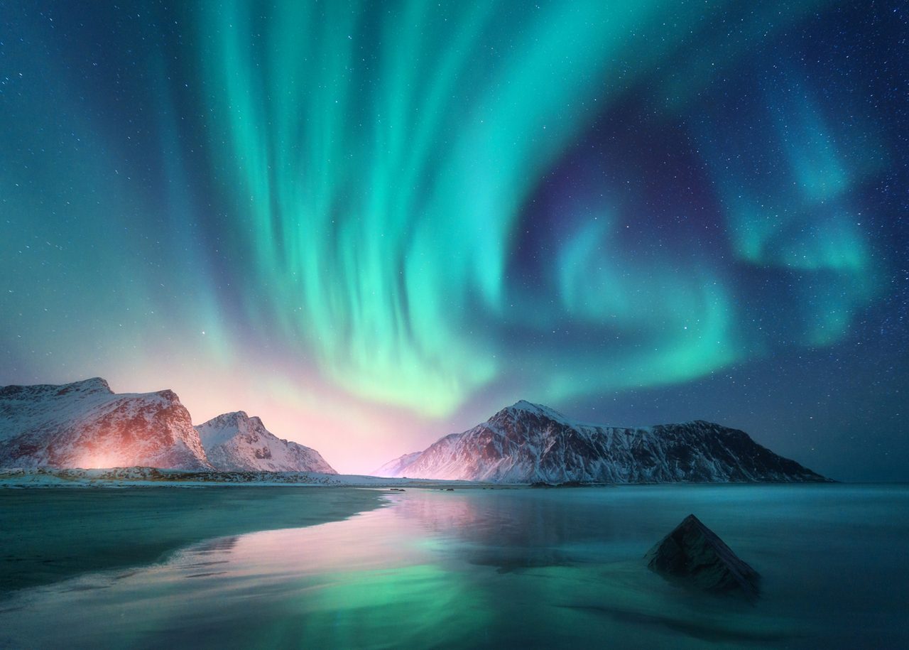 A breathtaking view of the Aurora Borealis, featuring vibrant green and teal light ribbons dancing across a starry night sky over the snow-capped mountains
