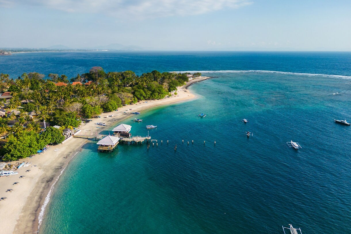 Aerial view of a Lombok beach showing white sand, lush palm trees, small waterfront pavilions and several boats floating in clear blue water along a curved shoreline.