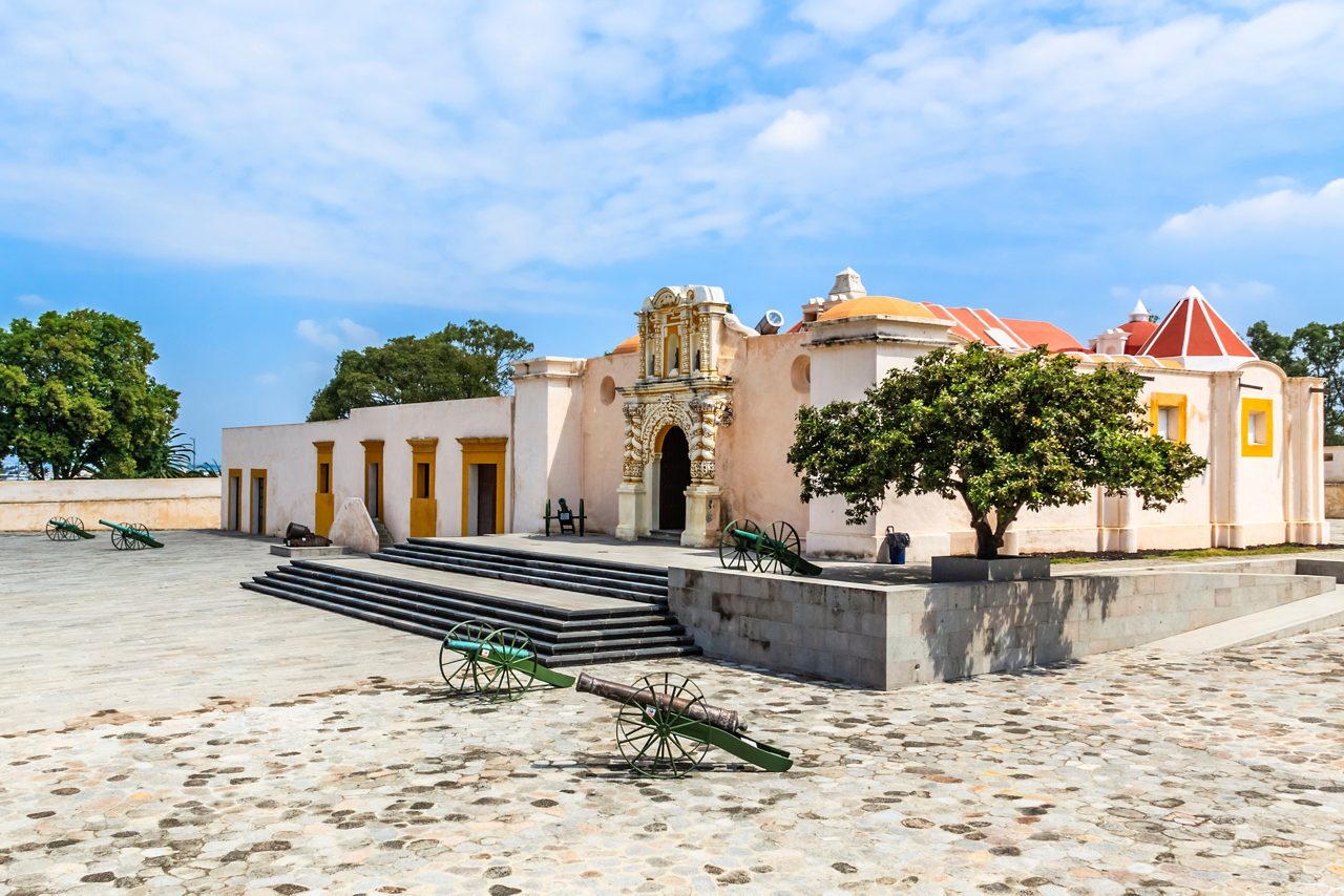 Historic fort in Loreto, Mexico, featuring a stone courtyard with cannons, a decorative arched entrance and pastel-colored colonial buildings under a clear sky.