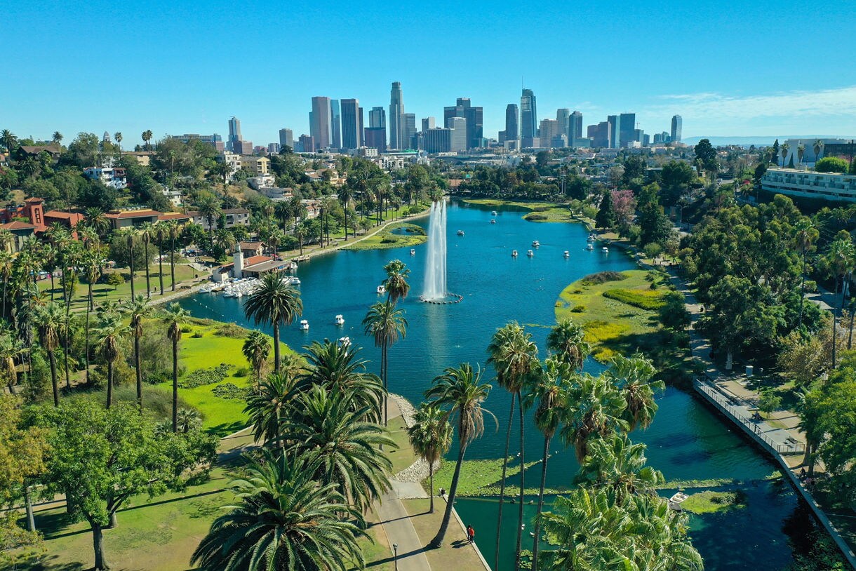 Aerial view of Echo Park Lake in Los Angeles with tall palm trees, a central fountain spraying water, small boats on the lake and the downtown skyline in the background.