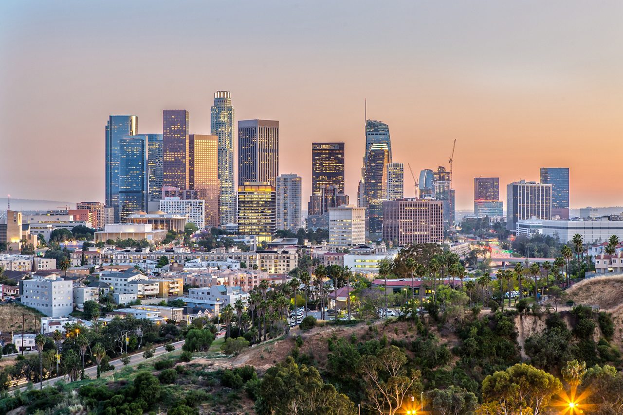 Cityscape at dusk, with a vibrant skyline and lush greenery in the foreground. The image features a mix of modern and older buildings, with a prominent road and streetlights. The sky transitions from blue to orange, creating a warm and inviting atmosphere. The overall scene exudes a sense of urban tranquility, with the city's energy and natural beauty harmoniously coexisting.