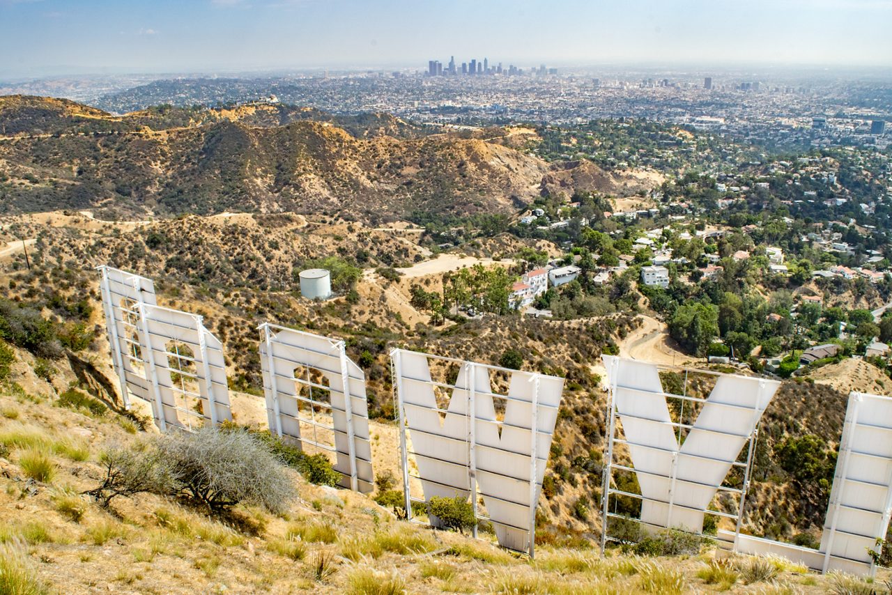 The iconic Hollywood sign, a symbol of the American film industry, stands proudly in the foreground of this image. The sign's white letters, arranged in a staggered formation, are supported by a metal frame and appear to be made of a translucent material. In the background, the sprawling city of Los Angeles stretches out, with its densely populated neighborhoods and towering skyscrapers. The hazy blue sky above adds a sense of depth and atmosphere to the image, while the dry, yellow grass in the foreground provides a sense of texture and context. 