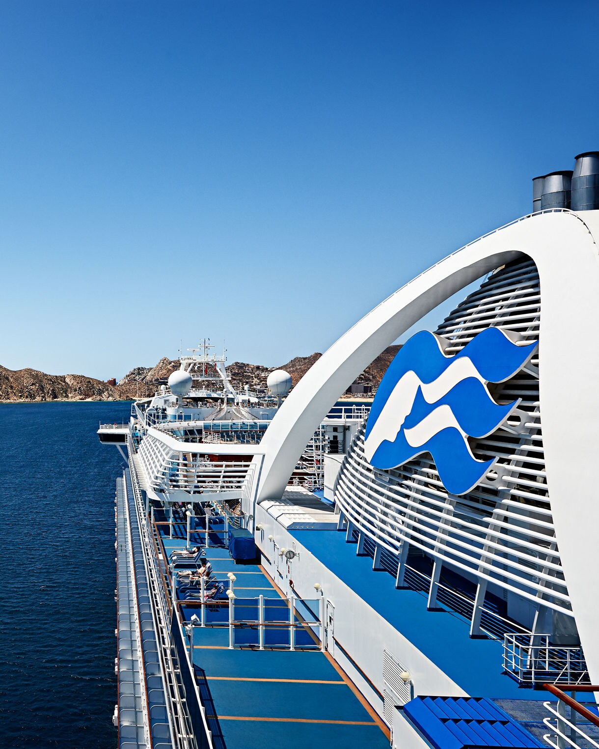 A side view of the Ruby Princess cruise ship with the iconic Princess Cruises logo, featuring a sunlit deck area and clear blue skies, with rocky coastal scenery in the background.