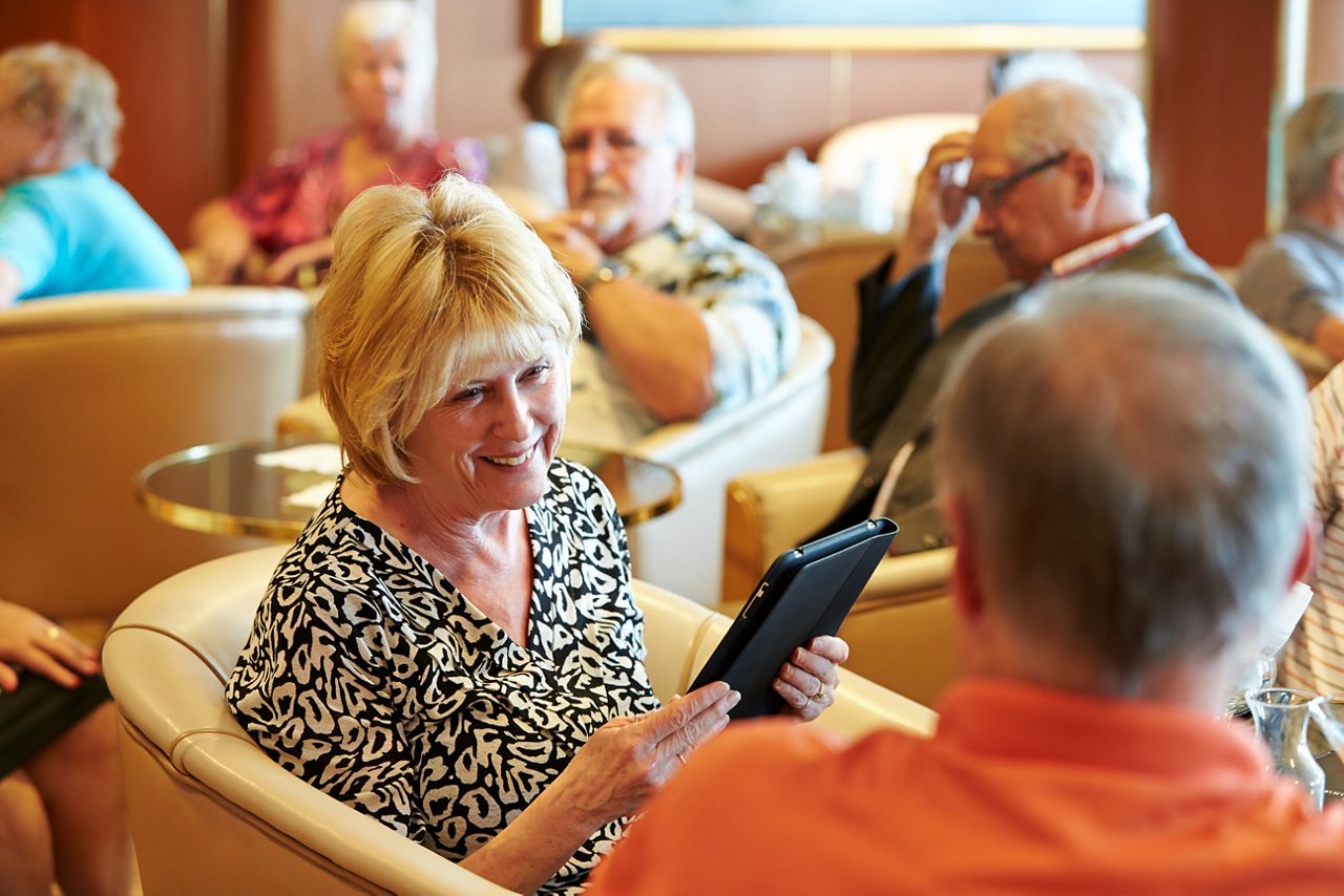 An older woman holds a tablet and smiles while seated in a ship lounge among other passengers.