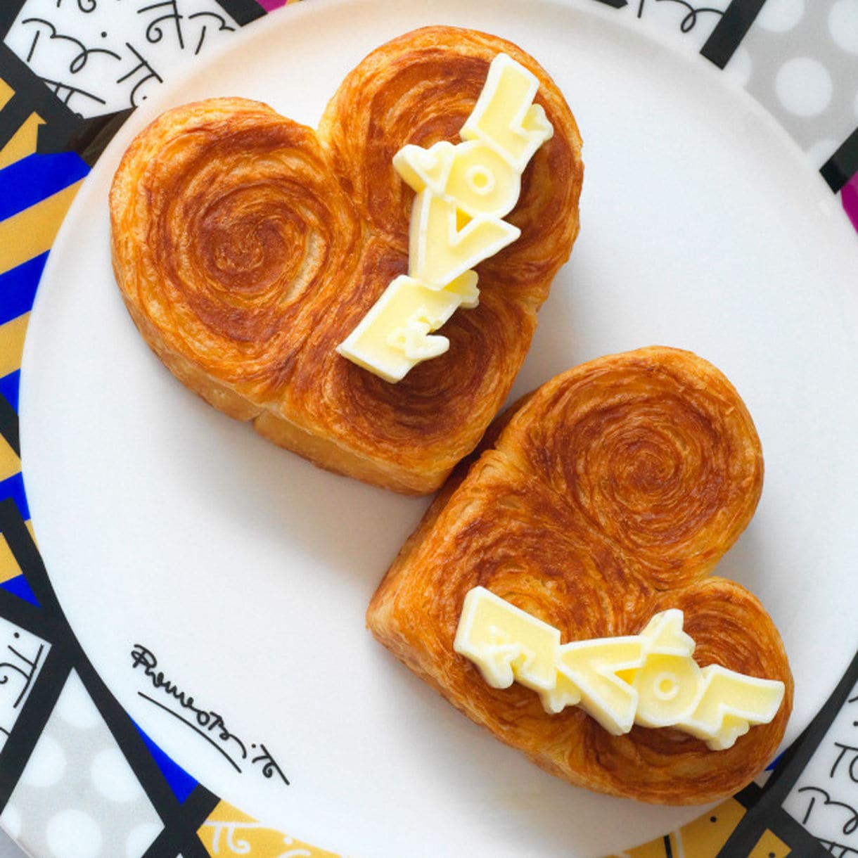 Two heart-shaped pastries with cream filling on a white plate, presented on a colorful patterned surface with Romero Britto inspired design.