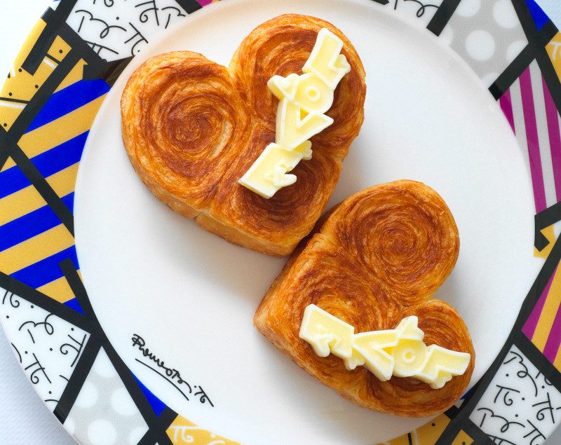 Two heart-shaped pastries with cream filling on a white plate, presented on a colorful patterned surface with Romero Britto inspired design.