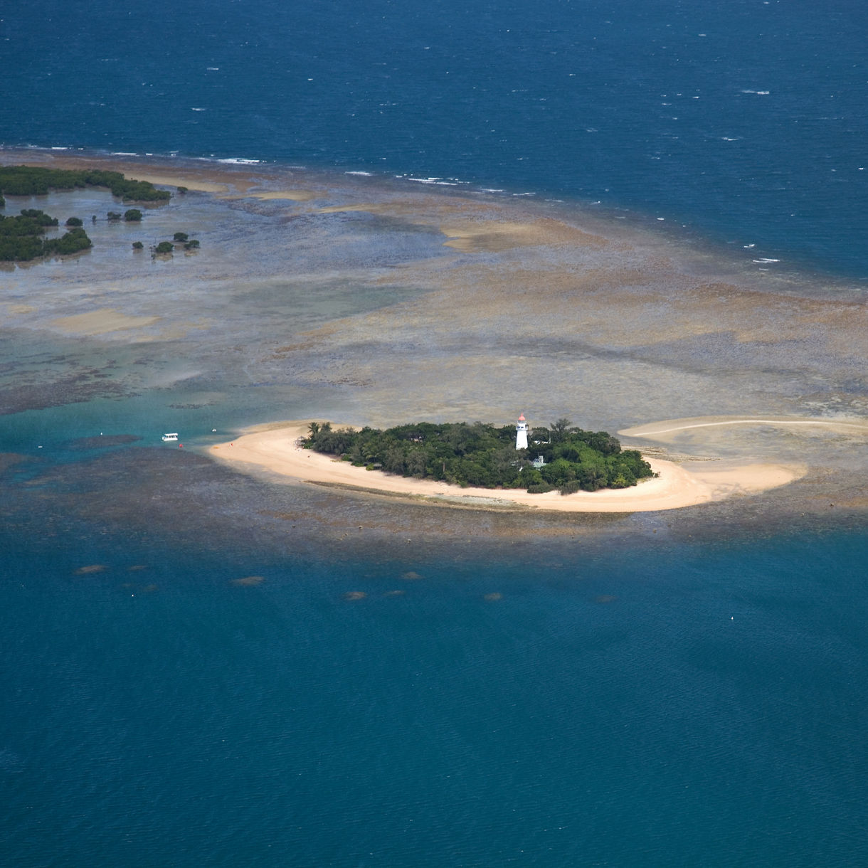 A small sandy island with a lighthouse and dense greenery, surrounded by shallow turquoise reef and deeper blue ocean.