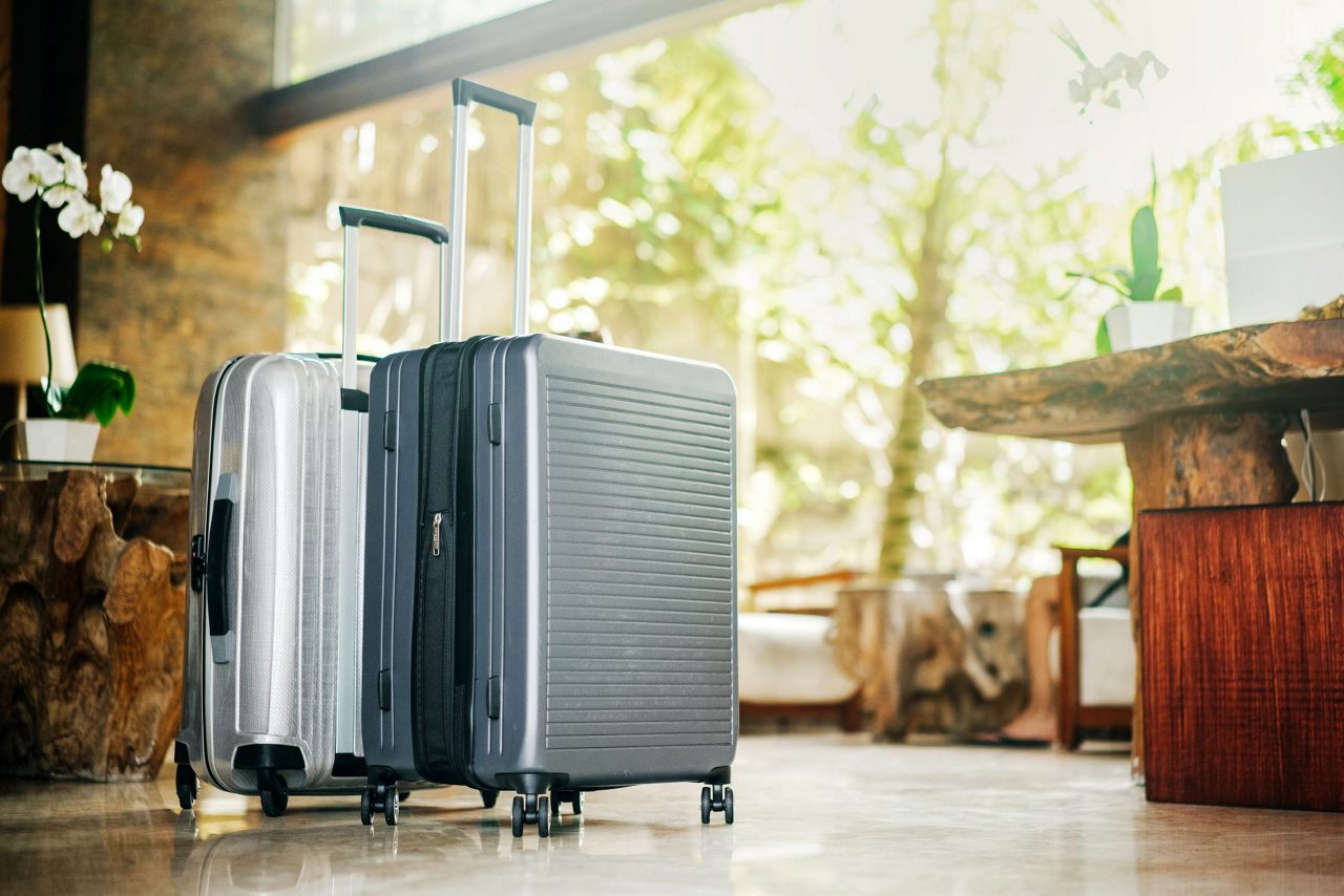 A silver rolling suitcase positioned in what appears to be a hotel or vacation rental entryway with wooden flooring.