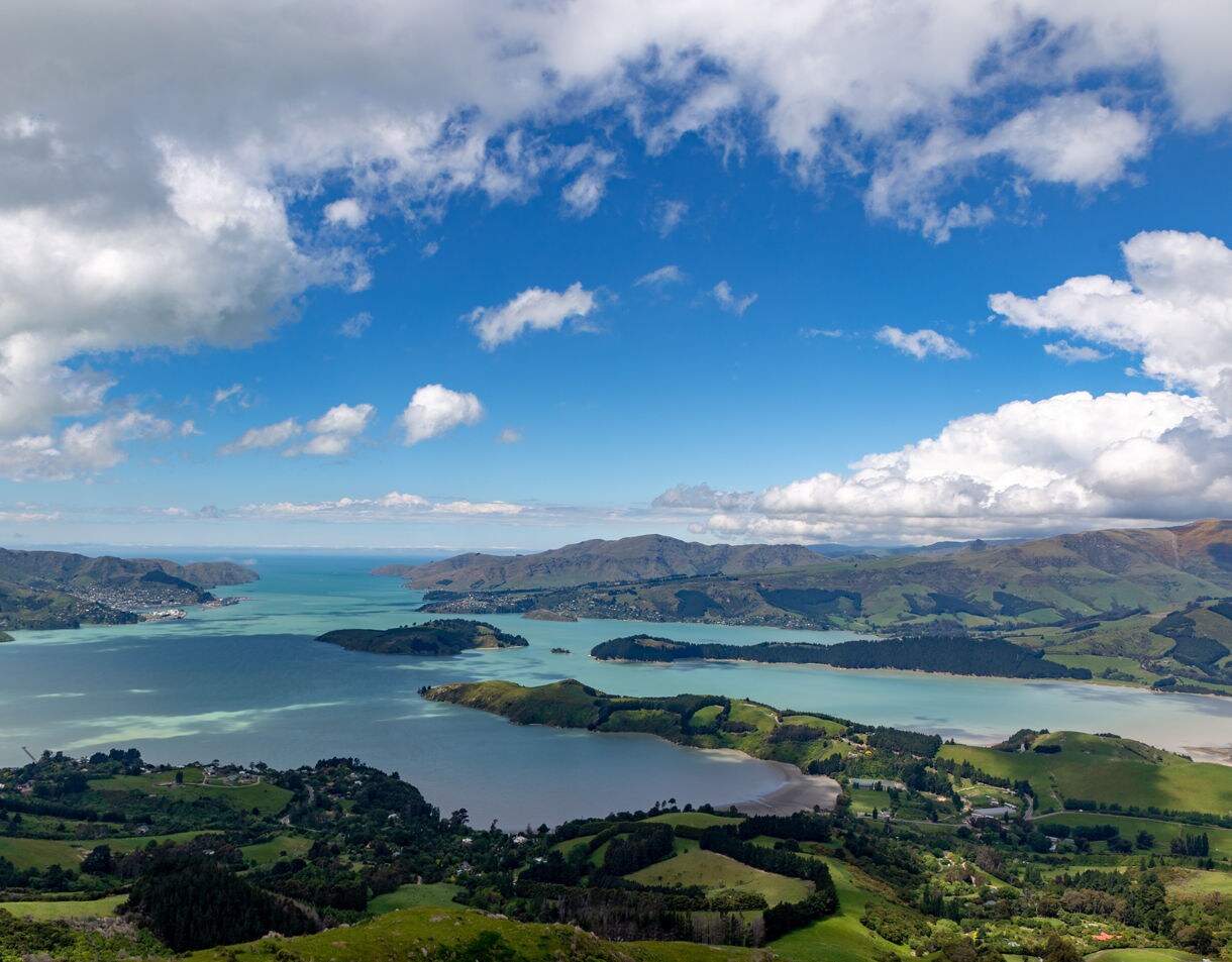 Aerial view of Lyttelton Harbour in New Zealand, showing vivid blue-green water surrounded by lush hills under a partly cloudy sky.