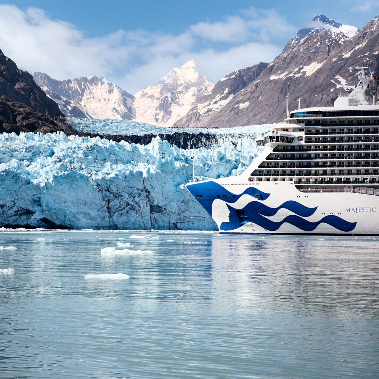 A large cruise ship is seen sailing close to a massive glacier, surrounded by icy waters and dramatic snow-capped mountains. The ship's bow features a distinctive blue wave design and the visible text 'MAJESTIC' on the hull. The scene captures the serene beauty of a remote, cold environment with floating ice chunks and clear reflections on the water.