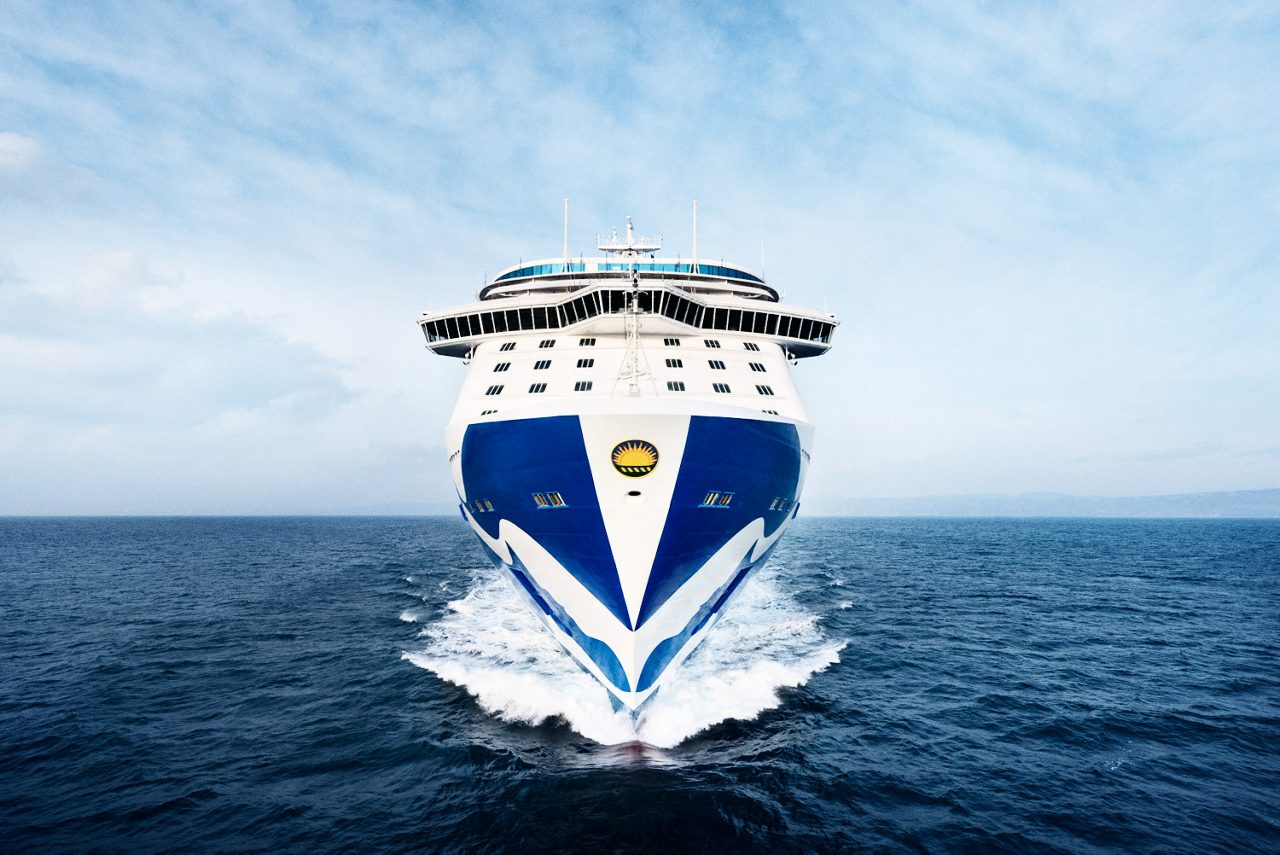 A front-facing view of a large Princess Cruises ship cutting through the ocean. The ship's bow, painted in blue and white with the iconic Princess Cruises logo, dominates the image as it moves steadily through the water under a bright blue sky. The vast sea stretches out behind the vessel.