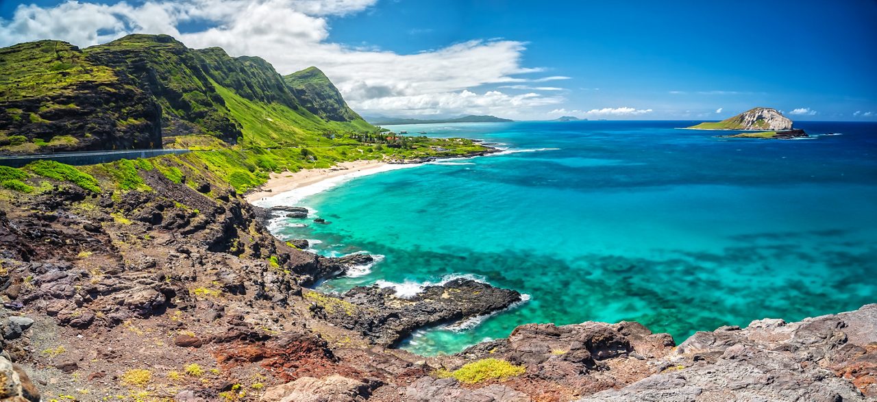 Panoramic view of Makapuʻu Point on Oahu with rocky cliffs, green hillsides, sandy beach and bright turquoise ocean stretching toward a small offshore island.