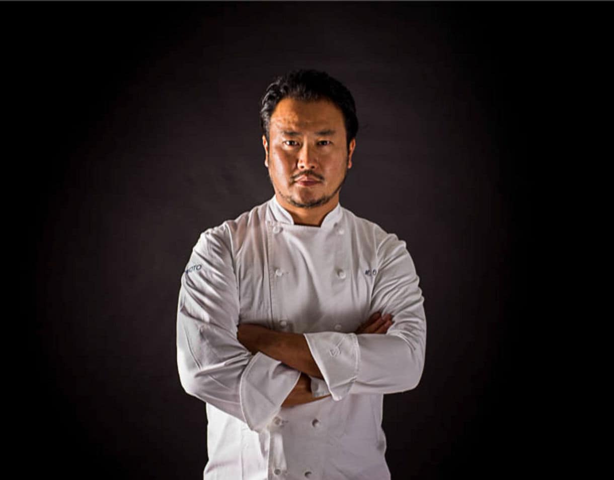 A Makoto Okuwa in traditional white uniform stands with crossed arms against a dark background, exuding confidence and professionalism.