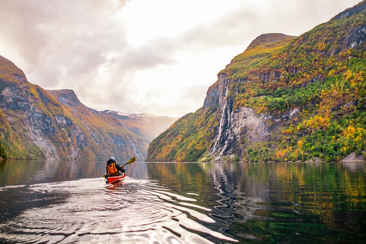 A person in a red kayak paddling through a calm Norwegian fjord surrounded by steep, forested cliffs and a distant waterfall on a cloudy day.