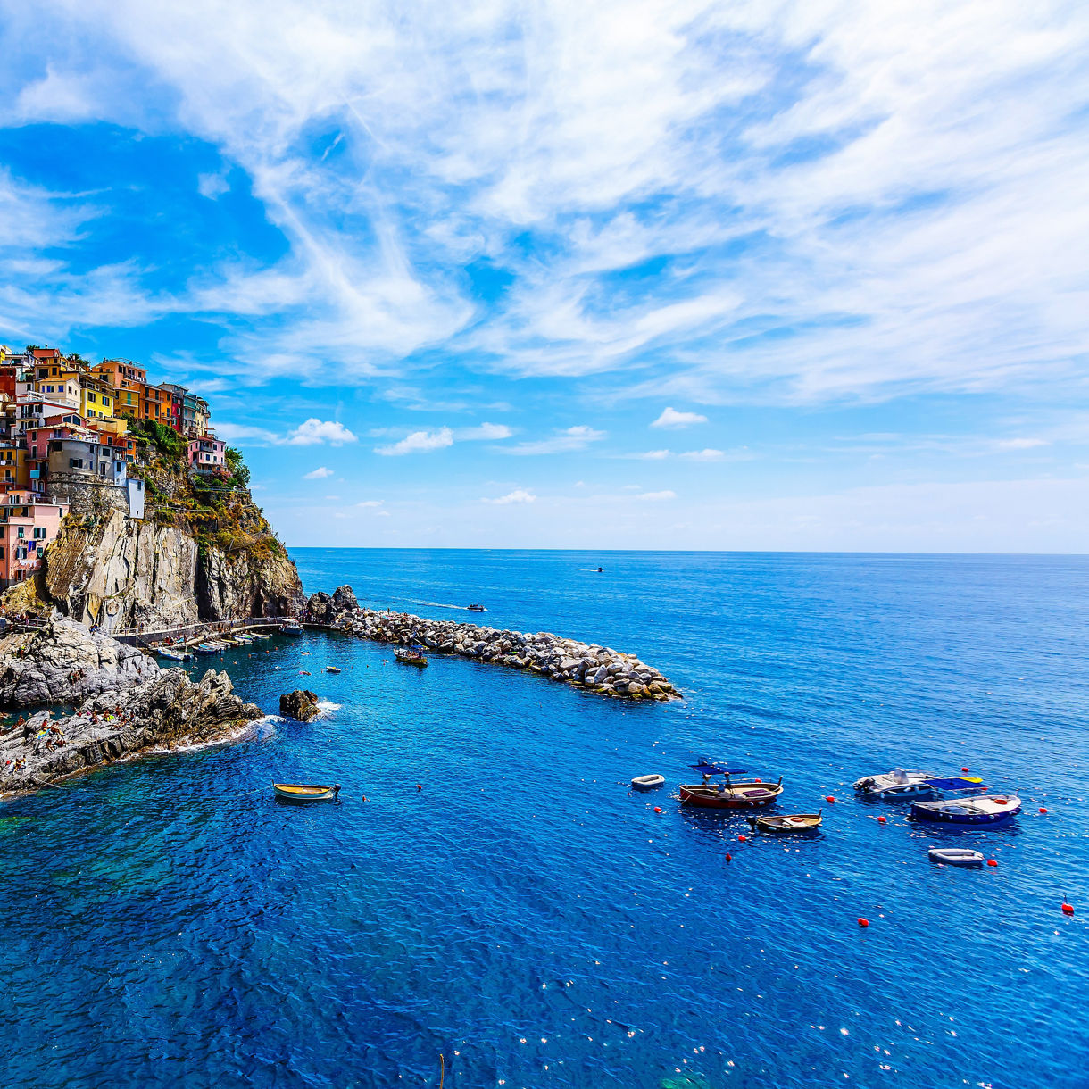 A coastal village in Cinque Terre with brightly painted houses stacked on rocky cliffs above deep blue water, small boats floating near the harbor.