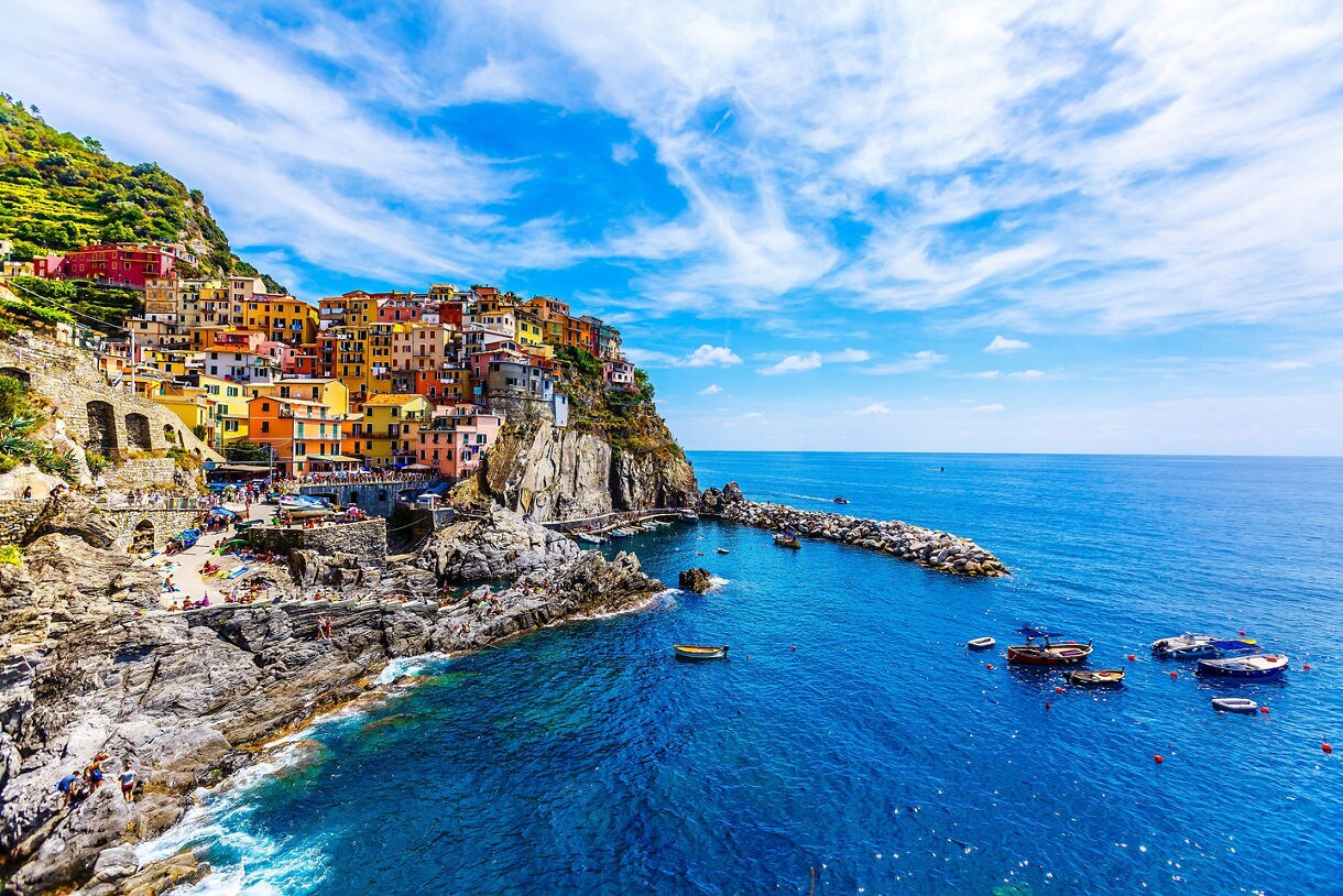A coastal village in Cinque Terre with brightly painted houses stacked on rocky cliffs above deep blue water, small boats floating near the harbor.