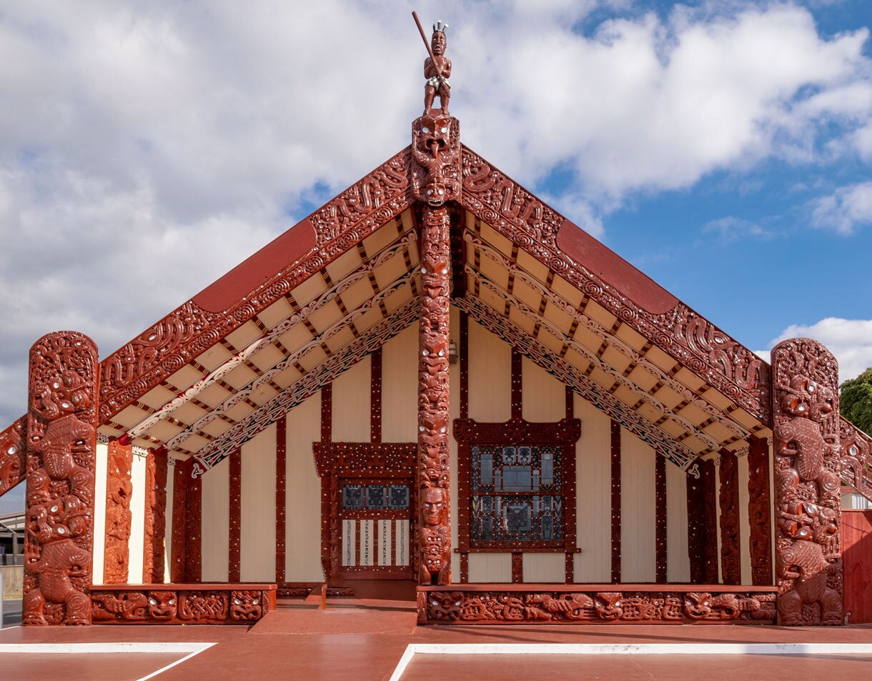 Traditional Māori meetinghouse with detailed red carvings and cream panels, featuring symbolic figures and patterns beneath a partly cloudy sky.