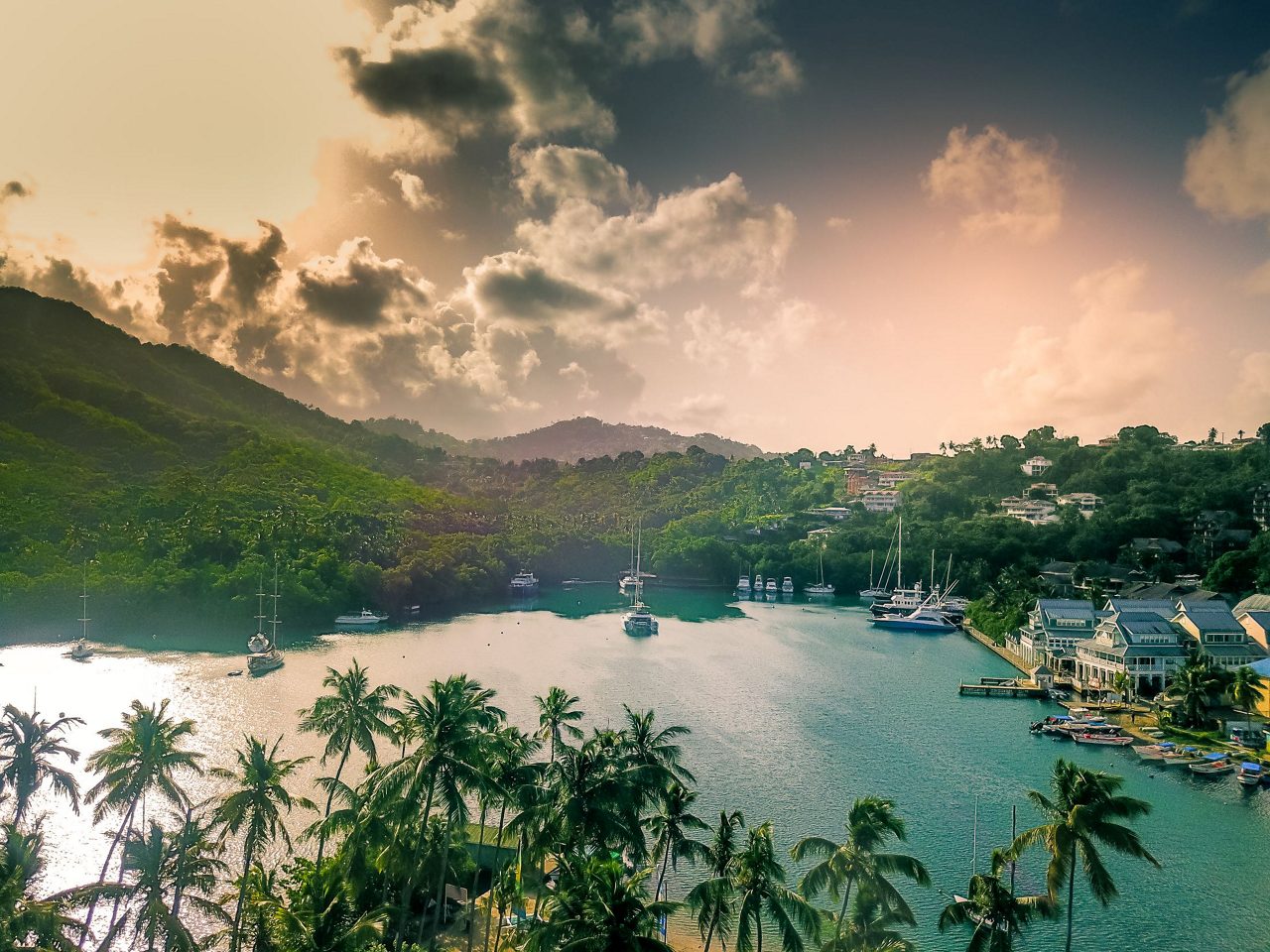 Scenic tropical bay at sunset with palm trees in foreground, sailboats anchored in calm waters, and green hills under golden sky in St. Lucia.