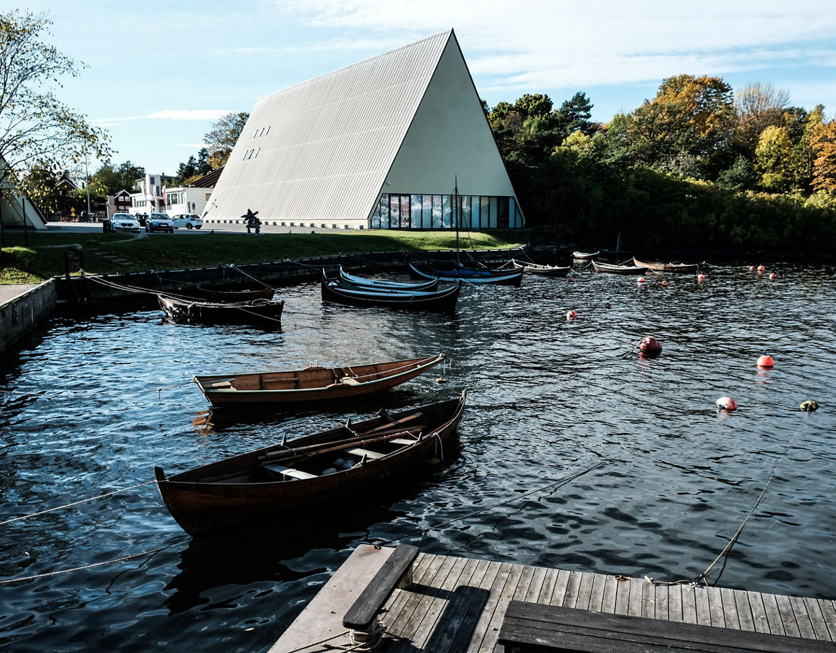 View of the Viking Ship Museum in Oslo, featuring a triangular modern building beside a harbor with small wooden boats floating on calm water.