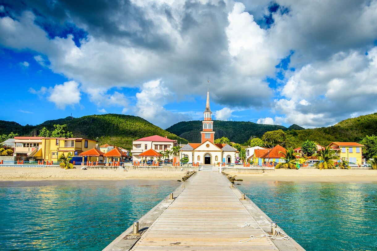 A scenic view of Anses-d’Arlet in Martinique featuring a wooden pier leading to a white church with a tall steeple, surrounded by colorful buildings, sandy beaches and green mountains under a bright blue sky with billowing clouds.