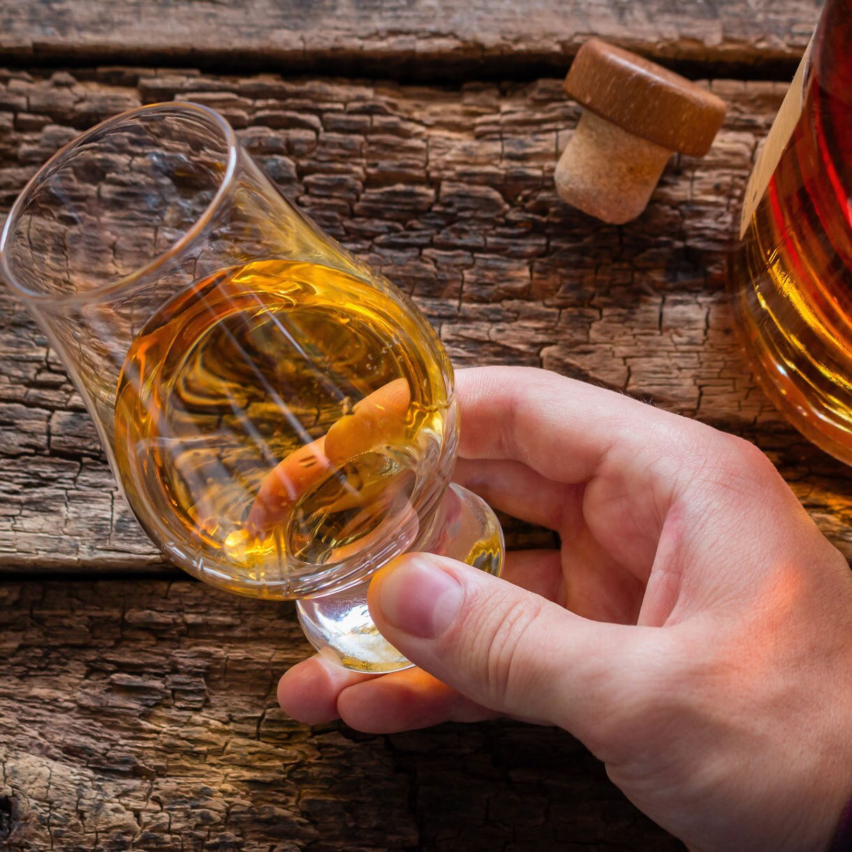 Close-up of a hand holding a glass of amber rum next to a cork and bottle on rustic wooden table in Martinique